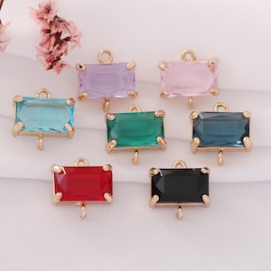 May include: Seven rectangular gemstone charms in various colors: light blue, purple, pink, green, dark blue, red, and black. Each charm has a gold-colored metal frame and a loop at the top for attaching to jewelry. The charms are displayed on a white fabric surface.
