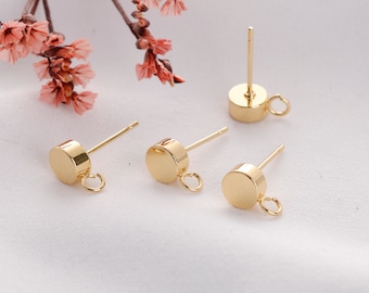 May include: Close-up of several gold-colored earring posts with a flat, round design. Each post has a small loop attached to the base and a straight post for insertion. The earrings are displayed on a white surface, with pink flowers in the background.