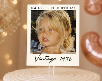 May include: A rectangular cake topper featuring a photograph of a young child with blonde hair. The text EMILY'S 30TH BIRTHDAY is printed at the top, and the text Vintage 1996 is printed at the bottom. The topper is mounted on two thin white sticks.
