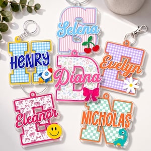 May include: A collection of six acrylic letter keychains featuring various patterns including blue gingham, pink stripes, floral prints, and checkered designs. Each keychain displays a personalized name in script or block text with small icons like a rocket, cherries, a daisy, a bow, a smiley face, and a dinosaur.
