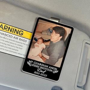 May include: A black rectangular car visor accessory with a photo of a man holding a baby. The text reads "MY GUARDIAN ANGEL RIDES SHOTGUN Dad 1955-2025" in white. The visor is attached to a gray car interior.
