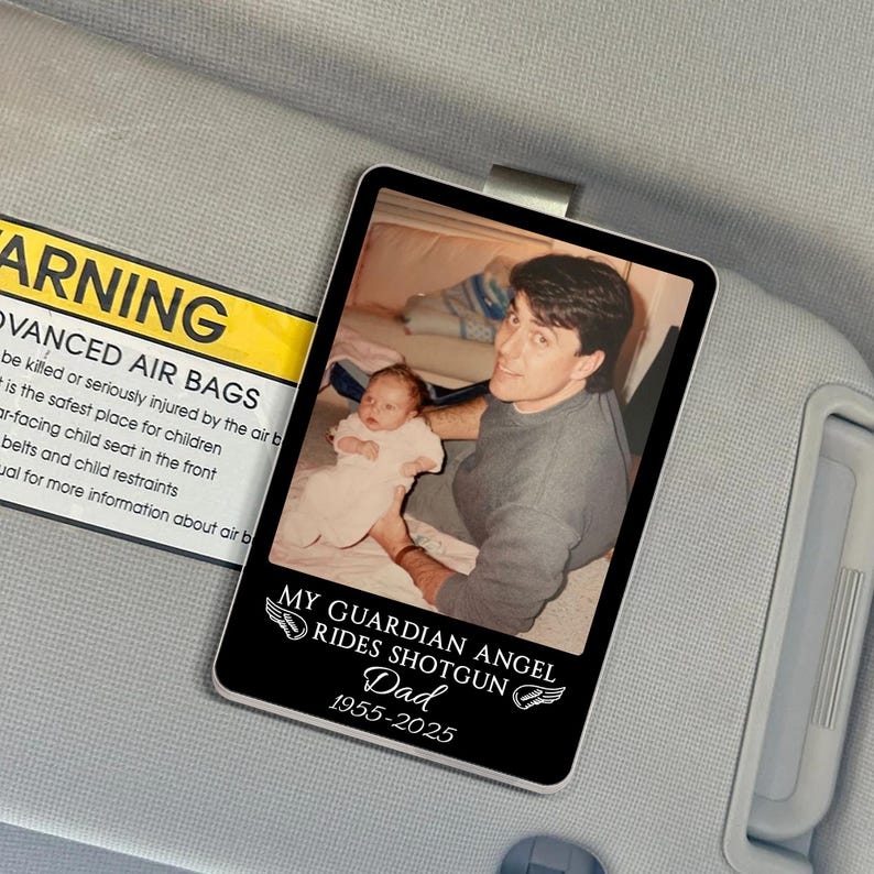 May include: A black rectangular car visor accessory with a photo of a man holding a baby. The text reads "MY GUARDIAN ANGEL RIDES SHOTGUN Dad 1955-2025" in white. The visor is attached to a gray car interior.