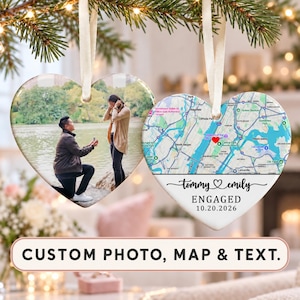 May include: Two heart-shaped ceramic ornaments hanging by cream ribbons. One ornament displays a photograph of a person proposing to another by a lake. The other ornament features a map of New York City with a red heart icon, the names Tommy and Emily, the word ENGAGED, and the date 10.20.2026.