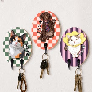 May include: Three oval wooden wall hooks featuring illustrations of pets. The left hook shows a calico cat labeled Daisy, the center shows a brown dog labeled Lola, and the right shows a white cat in a yellow cone labeled Jess. Each has black metal hooks.