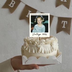 K&ouml;nnte beinhalten: Ein kleiner wei&szlig;er Kuchen mit ger&uuml;schtem Zuckerguss in einer wei&szlig;en Papierschale, gekr&ouml;nt von einer rechteckigen Karte mit dem Foto eines kleinen Kindes, dem Text HANNAH'S 30TH BIRTHDAY oben und dem handschriftlichen Text Vintage 1996 unten.