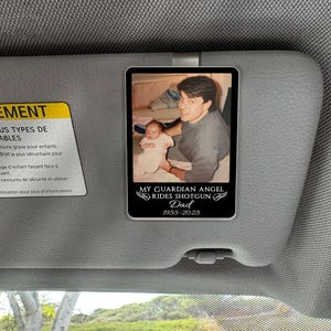 May include: A rectangular car visor accessory featuring a photograph of a man and a baby. The text "MY GUARDIAN ANGEL RIDES SHOTGUN Dad 1955-2025" is below the photo. The visor is attached to a car's interior.