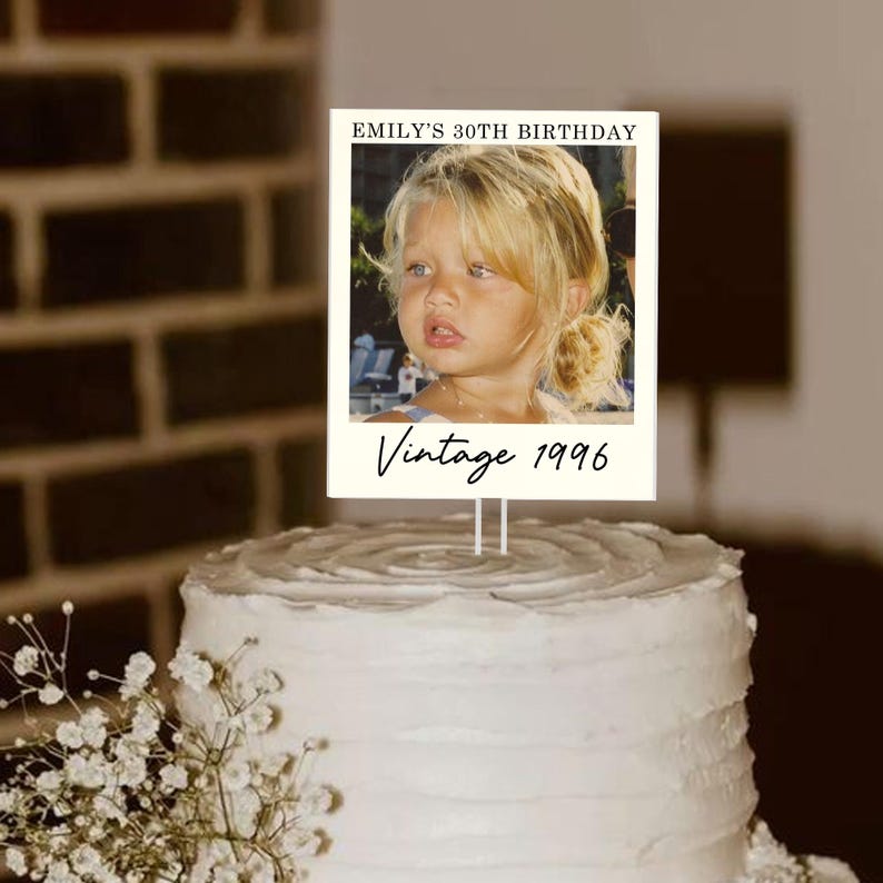 May include: A white cake with a decorative topper that reads "EMILY'S 30TH BIRTHDAY" and "Vintage 1996." The topper features a photo of a young girl. The cake is decorated with white frosting and small white flowers.