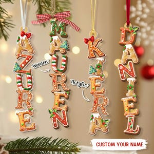 May include: Four vertical hanging ornaments featuring gingerbread-style letters spelling the names AZURE, MAUREEN, KAIERA, and DANIEL. Each letter is decorated with icing, holly, and small Christmas trees, suspended by ribbons.