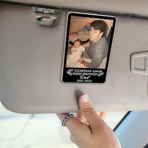 May include: A rectangular car visor accessory featuring a photograph of a man and a baby. The image is framed in black, with the text "MY GUARDIAN ANGEL RIDES SHOTGUN Dad 1955-2025" in white. The visor is gray.