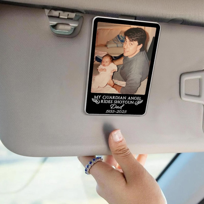 May include: A rectangular car visor accessory featuring a photograph of a man and a baby. The image is framed in black, with the text "MY GUARDIAN ANGEL RIDES SHOTGUN Dad 1955-2025" in white. The visor is gray.