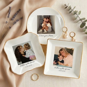 May include: Three white ceramic trinket dishes with gold rims, each featuring a photograph. One reads "I love you Mom," another "Mother of the Bride 12.20.2026," and the third "Mother of the Groom 10.09.2026." A gold ring and earrings are also present.