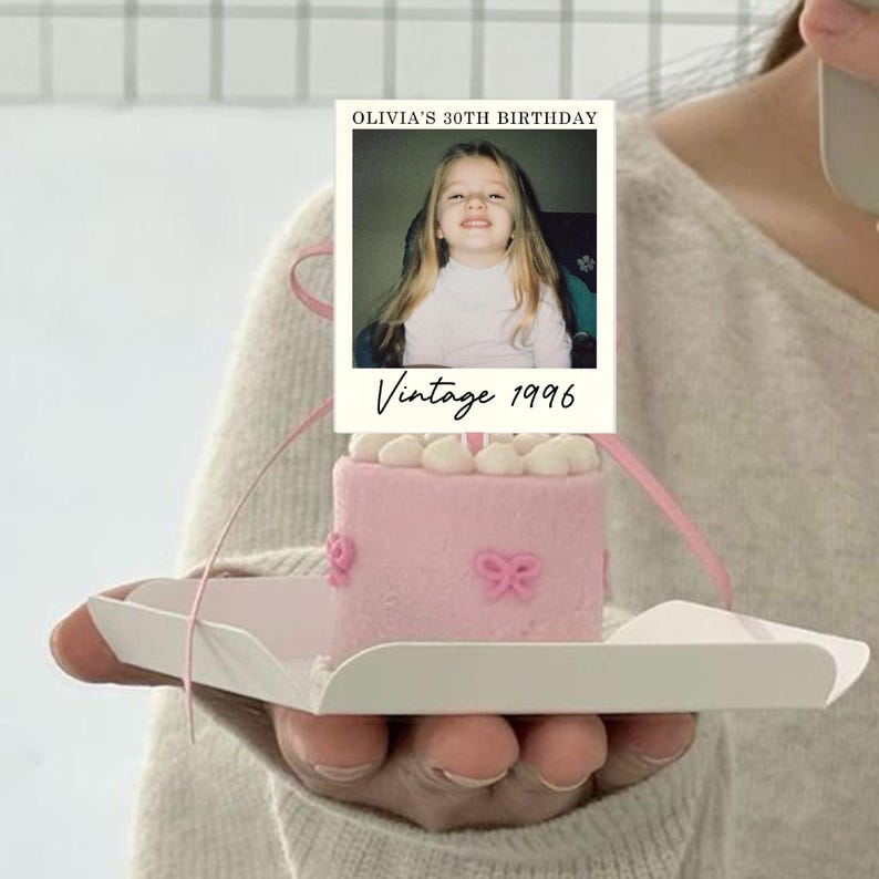 K&ouml;nnte beinhalten: Ein kleiner rosa Kuchen, verziert mit hellrosa Zuckerguss-Schleifen, gekr&ouml;nt von einer rechteckigen Karte mit dem Foto eines kleinen Kindes und dem Text OLIVIA'S 30TH BIRTHDAY sowie Vintage 1996, pr&auml;sentiert auf einem wei&szlig;en quadratischen Karton.