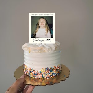 K&ouml;nnte beinhalten: Ein kleiner wei&szlig; glasierter Kuchen mit bunten Streuseln am Boden, gekr&ouml;nt von einer rechteckigen Karte mit dem Foto eines kleinen Kindes, dem Text OLIVIA'S 30TH BIRTHDAY und dem handschriftlichen Schriftzug Vintage 1996.