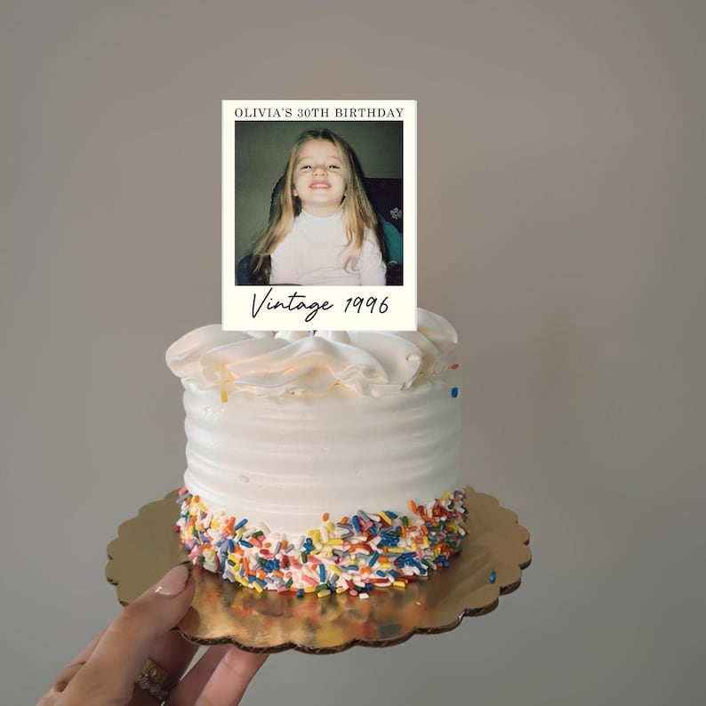 K&ouml;nnte beinhalten: Ein kleiner wei&szlig; glasierter Kuchen mit bunten Streuseln am Boden, gekr&ouml;nt von einer rechteckigen Karte mit dem Foto eines kleinen Kindes, dem Text OLIVIA'S 30TH BIRTHDAY und dem handschriftlichen Schriftzug Vintage 1996.