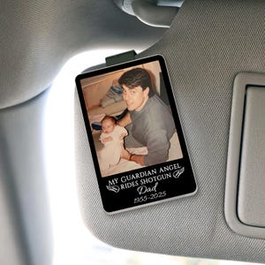 May include: A rectangular car visor ornament with a photo of a man and a baby. The ornament has the text "MY GUARDIAN ANGEL RIDES SHOTGUN Dad 1955-2025" in white. The ornament is clipped to a car visor.