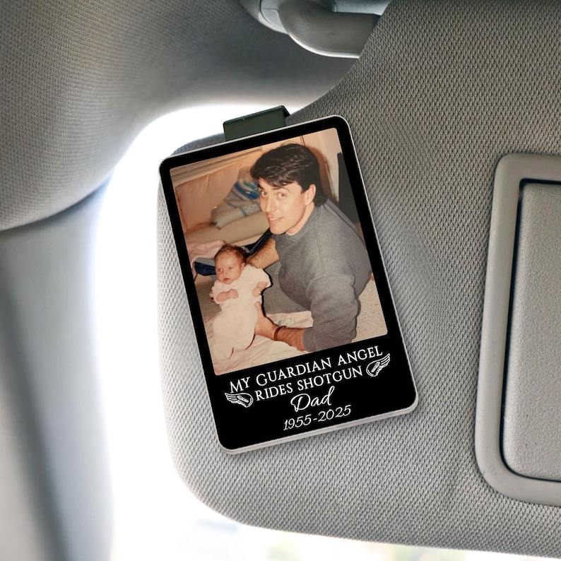 May include: A rectangular car visor ornament with a photo of a man and a baby. The ornament has the text "MY GUARDIAN ANGEL RIDES SHOTGUN Dad 1955-2025" in white. The ornament is clipped to a car visor.