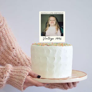 K&ouml;nnte beinhalten: Ein wei&szlig; glasierter Kuchen mit einem rechteckigen Tortenstecker, der ein Foto eines jungen Kindes zeigt. Der Stecker enth&auml;lt den Text OLIVIA'S 30TH BIRTHDAY oben und Vintage 1996 in einer Schreibschrift am unteren Rand des Fotorahmens.