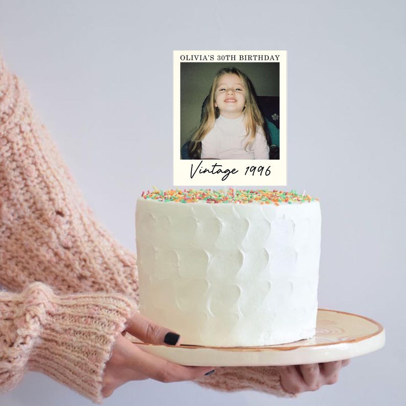 K&ouml;nnte beinhalten: Ein wei&szlig; glasierter Kuchen mit einem rechteckigen Tortenstecker, der ein Foto eines jungen Kindes zeigt. Der Stecker enth&auml;lt den Text OLIVIA'S 30TH BIRTHDAY oben und Vintage 1996 in einer Schreibschrift am unteren Rand des Fotorahmens.