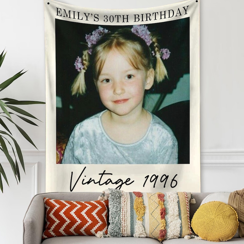 May include: A wall tapestry featuring a vintage photo of a young girl with the text "EMILY'S 30TH BIRTHDAY" at the top and "Vintage 1996" at the bottom. The photo is framed in white and hangs above a gray sofa with decorative pillows.