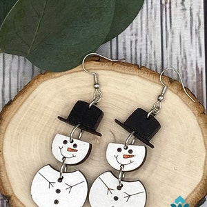 May include: A pair of wooden snowman earrings with black top hats and white faces. The earrings are hanging from silver hooks.