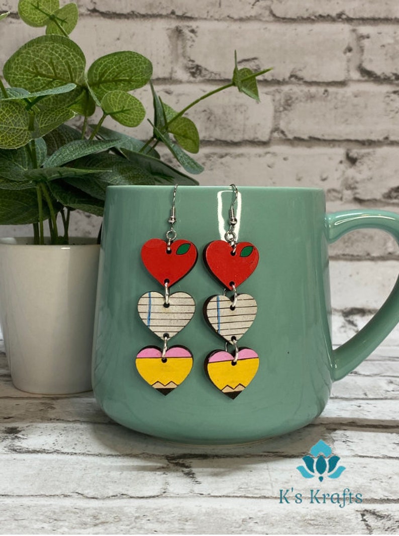 May include: A pair of dangle earrings with a school theme. Each earring features three heart-shaped charms: a red apple, a lined paper, and a yellow pencil. The earrings are made of wood and have silver-colored hooks.