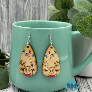 May include: A pair of wooden earrings with a reindeer design and colorful lights. The reindeer has a red nose and brown antlers. The earrings are hanging from a light blue mug.