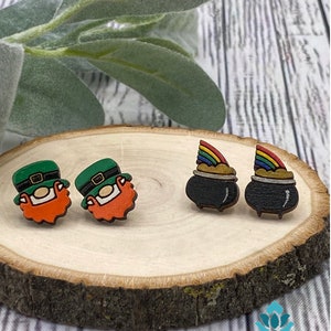 May include: A pair of wooden earrings featuring a leprechaun and a pot of gold. The leprechaun is wearing a green hat and has a red beard. The pot of gold has a rainbow on top. The earrings are displayed on a wooden surface.