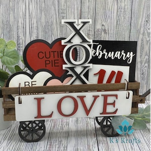 May include: A wooden wagon filled with Valentine's Day decorations. The wagon is painted brown and has black wheels. The decorations include a red heart with the words "Cutie Pie", a white heart with the word "Be", a pink heart, a black and white sign with the word "February", a red sign with the number "14", and a white sign with the word "LOVE" in red letters.