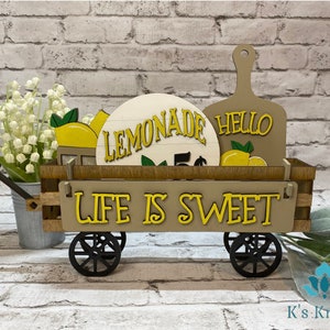 May include: A wooden wagon with black wheels, decorated with yellow and brown signs that say "Lemonade", "Hello", and "Life is Sweet". The wagon is filled with lemons and greenery.