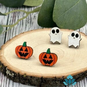 May include: A set of four Halloween-themed earrings. Two earrings feature a white ghost with black eyes and a smiling mouth. The other two earrings feature a jack-o'-lantern with an orange pumpkin and a black face.