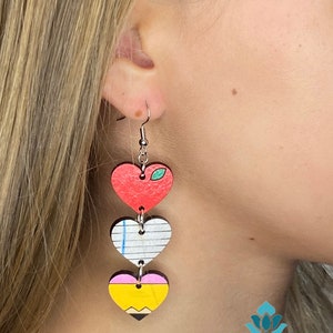 May include: Heart-shaped dangle earrings with a teacher theme. The earrings feature three stacked wooden hearts: a red apple, a lined paper, and a yellow pencil with a pink eraser and black tip. The earrings are suspended from silver-colored hooks.