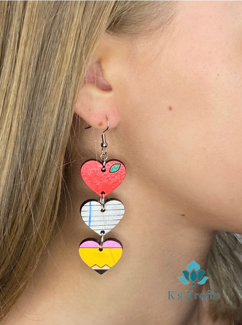 May include: Heart-shaped dangle earrings with a teacher theme. The earrings feature three stacked wooden hearts: a red apple, a lined paper, and a yellow pencil with a pink eraser and black tip. The earrings are suspended from silver-colored hooks.