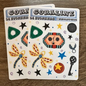 May include: Two sticker sheets with the word "CORALINE" and "14 STICKERS" printed on them. The stickers feature a variety of designs, including a dragonfly, stars, a key, and a gear shift. The stickers are in various colors, including green, orange, yellow, blue, and black.
