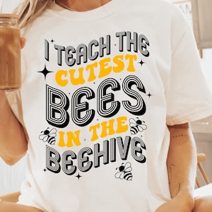 May include: White t-shirt with black and yellow text that reads "I teach the cutest bees in the beehive".