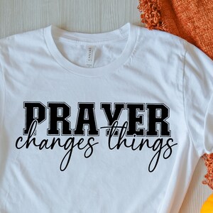 Peut inclure: T-shirt blanc avec le texte "PRAYER changes things" en lettres noires. Le t-shirt est présenté sur une surface en bois blanc avec des feuilles d'automne et des citrouilles.