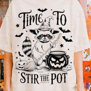 May include: Cream-colored t-shirt with a Halloween-themed graphic. The design features a raccoon wearing a witch hat, holding a broom, and stirring a cauldron. The text reads "Time To Stir The Pot."