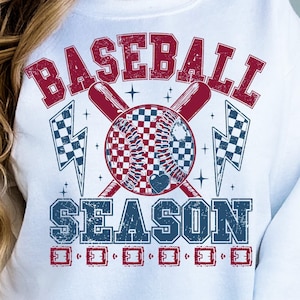 May include: White sweatshirt with a red and blue graphic design that says "Baseball Season". The graphic features a baseball with a checkered pattern, crossed bats, and lightning bolts.