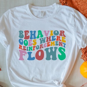 May include: White t-shirt featuring the text "Behaviour goes where reinforcement flows" in colourful, retro-style lettering. The shirt is laid out on a wooden surface with autumn leaves and pumpkins.