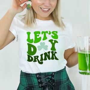 Puede incluir: Una camiseta blanca con texto verde que dice "Let's Day Drink" y un trébol verde. La persona lleva una falda de cuadros verdes y sostiene un vaso grande de líquido verde.
