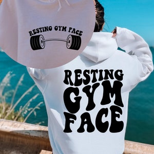 May include: White hoodie with black text that says "Resting Gym Face" in a retro font. The text is on the back of the hoodie. The front of the hoodie has a smaller version of the text with a barbell graphic.