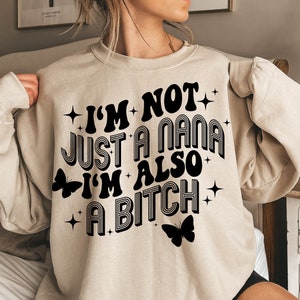 以下が含まれることがあります： ベージュ色のスウェットシャツ。黒色のレトロな文字で「I'M NOT JUST A NANA I'M ALSO A BITCH」と書かれています。デザインには黒い蝶のグラフィックと星のアクセントが含まれています。スウェットシャツは長袖です。