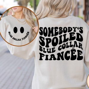 May include: A white sweatshirt with black text that reads "Somebody's Spoiled Blue Collar Fiancee" in a retro font. The back of the sweatshirt has a smiley face with the text "Blue Collar Fiancee".