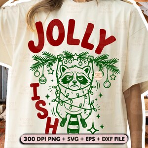 May include: A cream-coloured t-shirt featuring a graphic of a raccoon hanging from a green pine branch wrapped in string lights. The text reads JOLLY at the top and ISH at the bottom in red. The design includes ornaments and stars.