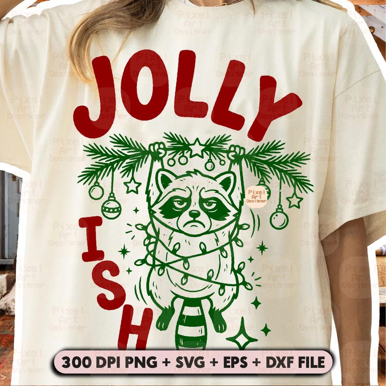 May include: A cream-coloured t-shirt featuring a graphic of a raccoon hanging from a green pine branch wrapped in string lights. The text reads JOLLY at the top and ISH at the bottom in red. The design includes ornaments and stars.