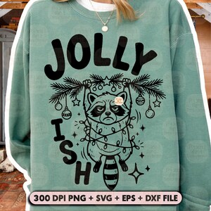 May include: A sage green crewneck sweatshirt featuring a black graphic of a raccoon hanging from a pine branch wrapped in string lights. The text reads JOLLY ISH in bold black letters. The bottom includes the text 300 DPI PNG + SVG + EPS + DXF FILE.