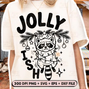 May include: A cream-coloured t-shirt featuring a black graphic of a raccoon tangled in Christmas lights, hanging from a pine branch with ornaments. The text reads JOLLY ISH. A bottom banner displays 300 DPI PNG + SVG + EPS + DXF FILE.