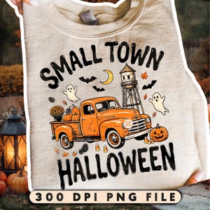 May include: A beige crewneck sweatshirt featuring a graphic print of an orange vintage pickup truck, a water tower, pumpkins, ghosts, bats, and autumn leaves. The text reads SMALL TOWN HALLOWEEN in black lettering. The fabric has a ribbed collar.
