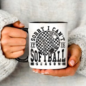May include: White ceramic mug with a black rim and handle. The mug features a black and white graphic of a softball with the text "Sorry I can't it is softball season".