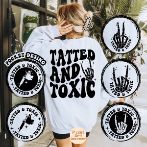 Puede incluir: Una sudadera blanca con el texto "TATTED AND TOXIC" en letras negras de estilo retro. La sudadera tiene cuatro diseños circulares en blanco y negro con el texto "TATTED & TOXIC" e imágenes de una pistola de tatuaje y una mano de esqueleto.