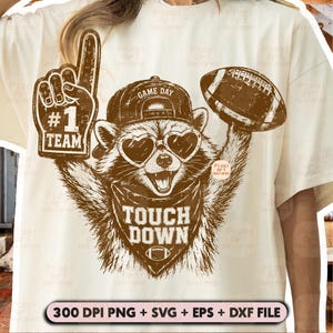 Peut inclure: Un t-shirt crème avec un motif marron représentant un raton laveur portant une casquette et des lunettes en forme de cœur. Le raton laveur tient un doigt en mousse #1 TEAM et un ballon de football. Texte : TOUCH DOWN et 300 DPI PNG + SVG + EPS + DXF FILE.